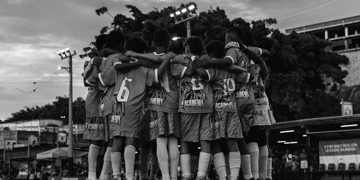 A black and white photo of a youth soccer team huddling on the field during dusk.